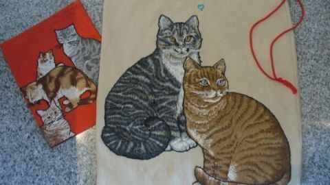 Eine Stofftasche zeigt zwei sitzende Katzen, eine grau und eine orange, in einer gemütlichen Umgebung, umgeben von einer weiteren roten Tasche.