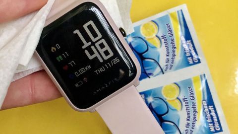 Eine Hand hält ein Tuch, um eine Armbanduhr mit Kautschukband zu reinigen, während Päckchen für Kunststoffpflege im Hintergrund liegen.