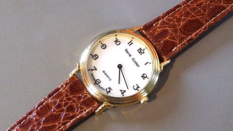 Eine goldene Armbanduhr mit braunem Lederarmband liegt auf einer grauen Oberfläche und zeigt die aktuelle Uhrzeit an.