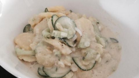 Kartoffelsalat mit Gurken liegt in einer weißen Schüssel; die Zutaten sind mit einer cremigen Sauce vermischt.