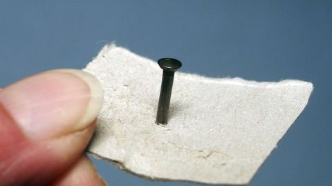 Ein Nagel steht senkrecht auf einem Stück Papier, während eine Hand ihn festhält, bereit zum einfachen Einschlagen.