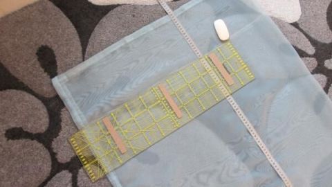 Ein transparenter Gardinenschal liegt flach auf einem gemusterten Teppich, während ein Lineal zum Messen daneben positioniert ist.