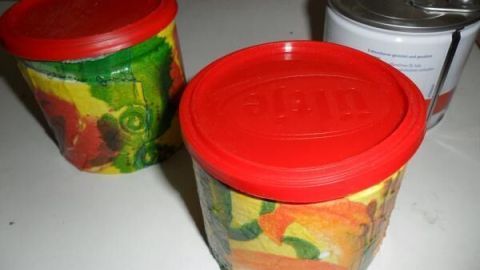 Zwei bunte Erdnussdosen mit roten Deckeln stehen dekorativ auf einem Tisch. Eine silberne Dose ist im Hintergrund.