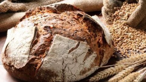 Ein knuspriges Brot mit dunkler Kruste liegt auf einem Holztisch, umgeben von Getreideähren und losem Weizen auf Jutestoff.