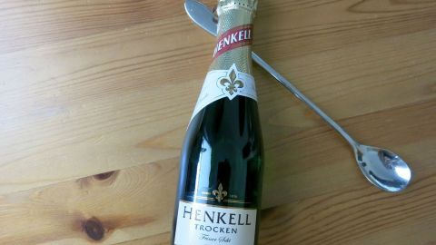 Ein Silberlöffel steckt in einer geöffneten Henkell Sektflasche, die auf einem Holztisch liegt.