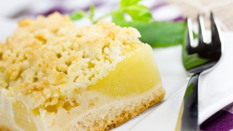 Ein Stück Kuchen mit Streusel liegt auf einem weißen Teller, daneben eine Gabel. Im Hintergrund sind grüne Blätter zu sehen.