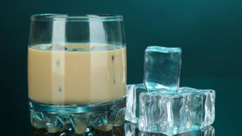 Ein Glas Karamelllikör steht auf einem Tisch neben einem Eiswürfel, vor einem dunklen, glatten Hintergrund.