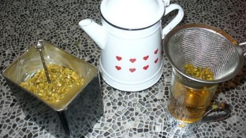 Ein Teesieb mit Misteltee über einem Glas, neben einer weißen Teekanne mit roten Herzen und einem Behälter mit getrockneten Kräutern.