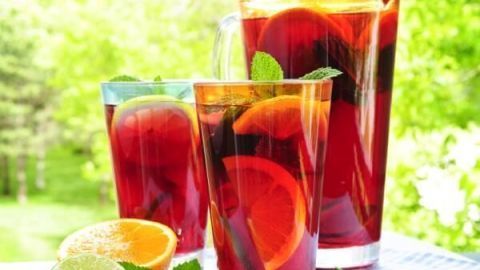 Gläser und ein Krug voller roter Trauben-Limonade mit Orangenscheiben und Minzblättern stehen auf einer Decke im Garten.