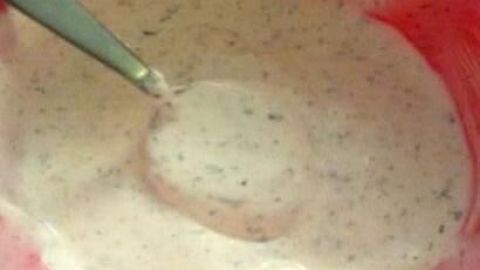 Ein cremiger Schmand-Knoblauch-Dip in einer roten Schüssel wird mit einem Löffel gut verrührt, um eine einheitliche Konsistenz zu erreichen.