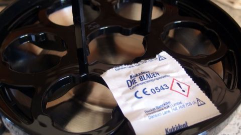 Eine schwarze Heizplatte eines elektrischen Eierkochers liegt auf einer Küchenoberfläche mit einem Sicherheitsetikett.