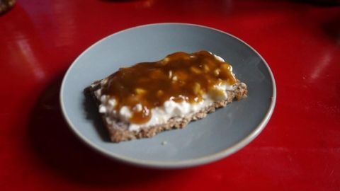 Eine Scheibe Brot ist mit Rhabarber-Aprikosen-Marmelade und Quark belegt und steht auf einem blauen Teller auf einem roten Tisch.