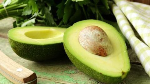 Avocadohälfte mit Kern liegt auf einem Holztisch neben einem Messer und einem Bündel Petersilie, im Hintergrund ein Geschirrtuch.