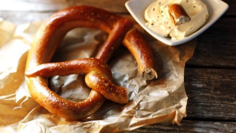 Ein frisch gebackenes Brezel liegt auf braunem Papier, neben einem Schälchen mit cremigem Obazda auf einem Holztisch.