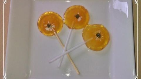 Drei Orangen-Lutscher mit Gänseblümchen liegen auf einem weißen Teller, umrahmt von einer blumigen Verzierung.