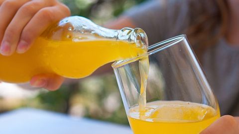 Eine Hand gießt ein orangefarbenes Getränk aus einer Flasche in ein Glas, im Hintergrund unscharfe grüne Natur.