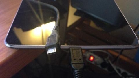 Ein Micro USB-Kabel steckt schief in einem Tablet, das auf einem Tisch liegt. Im Hintergrund sind Steckdosen zu sehen.