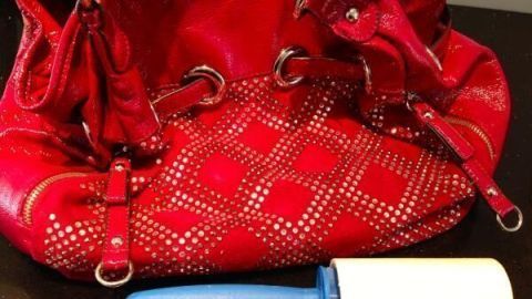 Eine rote Handtasche liegt auf einer Oberfläche neben einem blauen Fusselroller, bereit zur Reinigung.