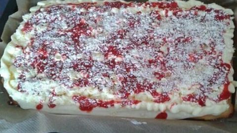 Rechteckiger Spaghettikuchen mit Sahne, Erdbeersoße und Kokosraspeln auf Backpapier in einer Küche präsentiert.