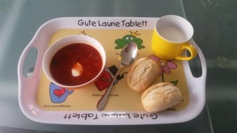 Ein Esstablett mit der Aufschrift "Gute Laune Tablett" hält Suppe, Brötchen, Löffel und eine gelbe Tasse mit Milch.