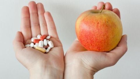 Eine Hand zeigt eine Auswahl an Nahrungsergänzungsmitteln, während die andere Hand einen frischen Apfel hält, was einen Vergleich von Vitaminpräparaten und natürlichen Lebensmitteln darstellt.