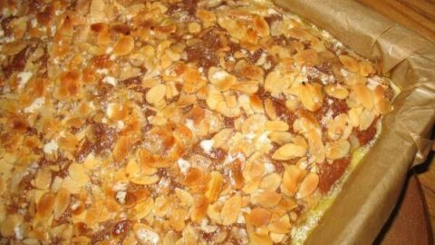 Eine goldbraune Buttermilch-Blechkuchen mit knuspriger Mandelkruste liegt in einer Backform auf einem Holzuntergrund.
