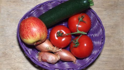Ein geflochtener Korb enthält frisches Obst und Gemüse, darunter Äpfel, Tomaten und Zucchini, auf einer Holzoberfläche.