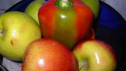 Eine grüne Paprika liegt in einer Schüssel umgeben von Äpfeln, während sie nachreifen soll, um ihre Farbe zu ändern.