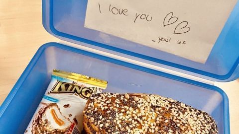 In einer blauen Brotdose steht ein Brötchen, neben dem ein Zettel mit liebevollen Botschaften und Snacks liegt.