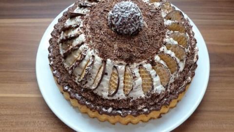 Eine Torte mit Schaumküssen, verziert mit Waffeln und Schokoraspeln, steht auf einem weißen Teller auf einem Holztisch.