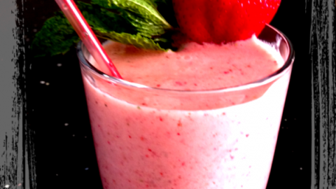 Ein Erdbeer-Smoothie in einem Glas mit Strohalm. Eine frische Erdbeere und Minze garnieren das Getränk auf einem dunklen Tisch.