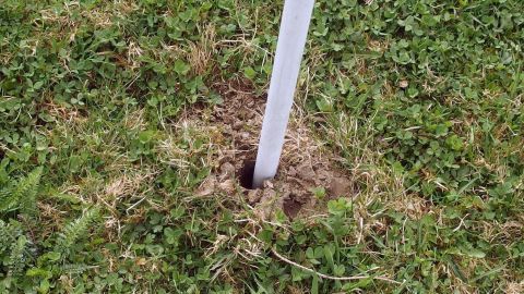 Ein Pfahl steht in einem Erdloch auf grünem Gras, möglicherweise zur Vertreibung von Wühlmäusen im Garten.