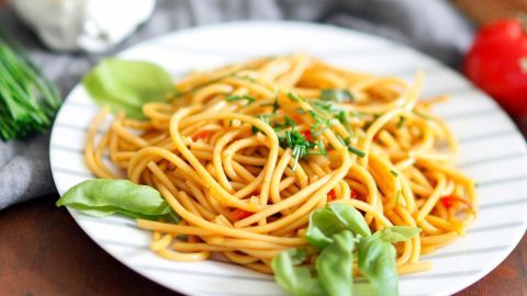 Ein Teller asiatischer Spaghettisalat liegt auf einem Holztisch, während frische Basilikumblätter und Gemüse um ihn herumliegen.