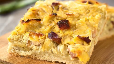 Ein frisch gebackener Zwiebelkuchen liegt auf einem Holzbrett, köstlich und leicht knusprig, umgeben von grünen Zutaten.