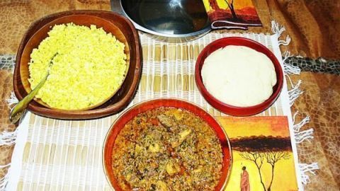 Egusi-Suppe mit Fleisch und Pilzen in einer roten Schüssel neben gelbem Reis und weißem Teig auf Bambusmatte serviert.
