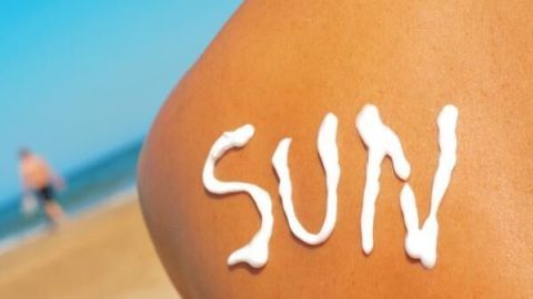 Ein mit Sonnencreme bedeckter Rücken zeigt das Wort "SUN" am Strand; verschwommener Mann läuft am Wasser entlang.