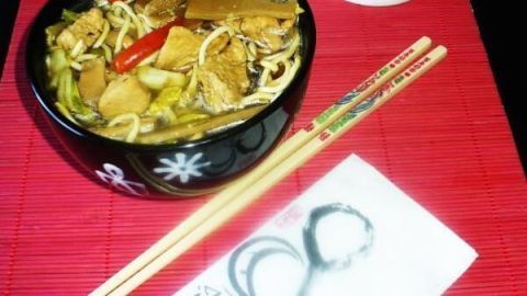 In einer Schüssel mit scharfer chinesischer Hühnernudelsuppe liegen frische Zutaten, während Glücks-Katze im Hintergrund steht.