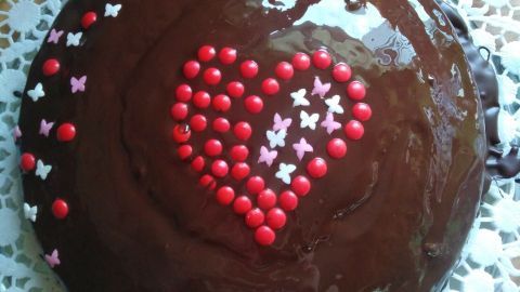 Ein großer Schokokuchen, glasiert und mit bunten Zuckerstreuseln in Herzform dekoriert, steht auf einem weißen Tisch.