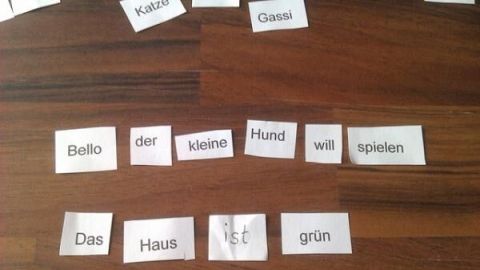 Papierstreifen mit Wörtern wie "Bello", "Hund", "Haus" und "grün" bilden Sätze auf einem Holztisch, üben Satzbildung.