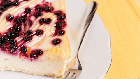 Eine Stück Heidelbeer-Schmand-Torte liegt auf einem weißen Teller, garniert mit schwarzen Beeren und einer Gabel daneben.