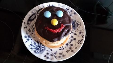 Ein mit Schokolade überzogener Schaumkuss zeigt ein Gesicht mit bunten Smarties als Augen und Nase, auf weißem, blumigem Teller.