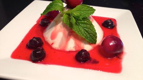 Eine Panna Cotta in Vanilleweiß sitzt auf roter Erdbeersauce, garniert mit frischen Minzblättern und umgeben von Kirschen.
