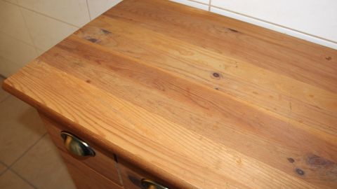 Ein Holzschrank mit einer leicht beschädigten, unlackierten Oberfläche steht in einer hellen Küche.