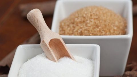 Zucker in zwei Schalen: Vorne weißer Zucker mit Holzschaufel, hinten brauner Zucker; Nahaufnahme auf Holztisch.