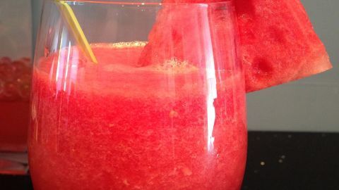 Ein erfrischender Wassermelonen-Drink steht in einem Glas mit einem gelben Strohhalm und einem Stück Wassermelone oben drauf.