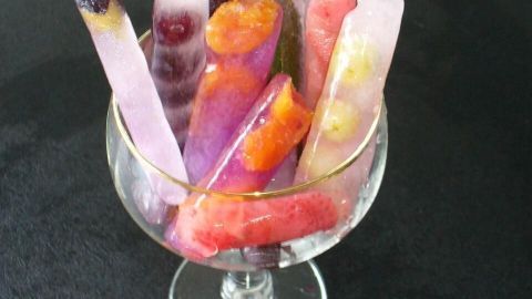 Eisstäbchen aus frischen Früchten stehen in einer Schale, umgeben von bunten Fruchtstücken auf einem dunklen Hintergrund.