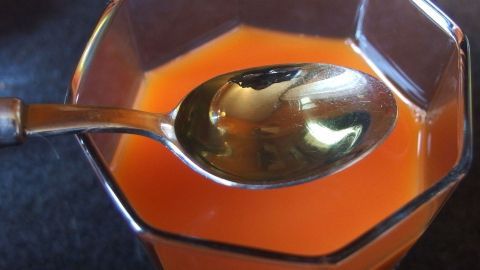 Karottensaft mit Öl trinken - dadurch kann der Körper das Vitamin A aufnehmen, ansonsten würde dies verloren gehen. Ein Glas Karottensaft steht auf einem Tisch, während ein Löffel schräg im Saft steckt und reflektiert.