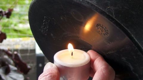 Ein Blumentopf wird mit einer Hand gehalten, während ein Teelicht darin brennt; eine ruhige Gartenatmosphäre umgibt die Szene.