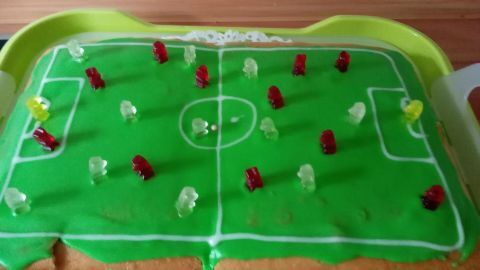 Ein Fußballkuchen liegt auf einer grünen Fläche, dekoriert mit bunten Figuren, die ein Spiel darstellen.