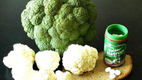 Brokkoli und Blumenkohl liegen auf Schwarzbrot neben einem geöffneten Kaiser Natron Behälter auf schwarzem Untergrund.